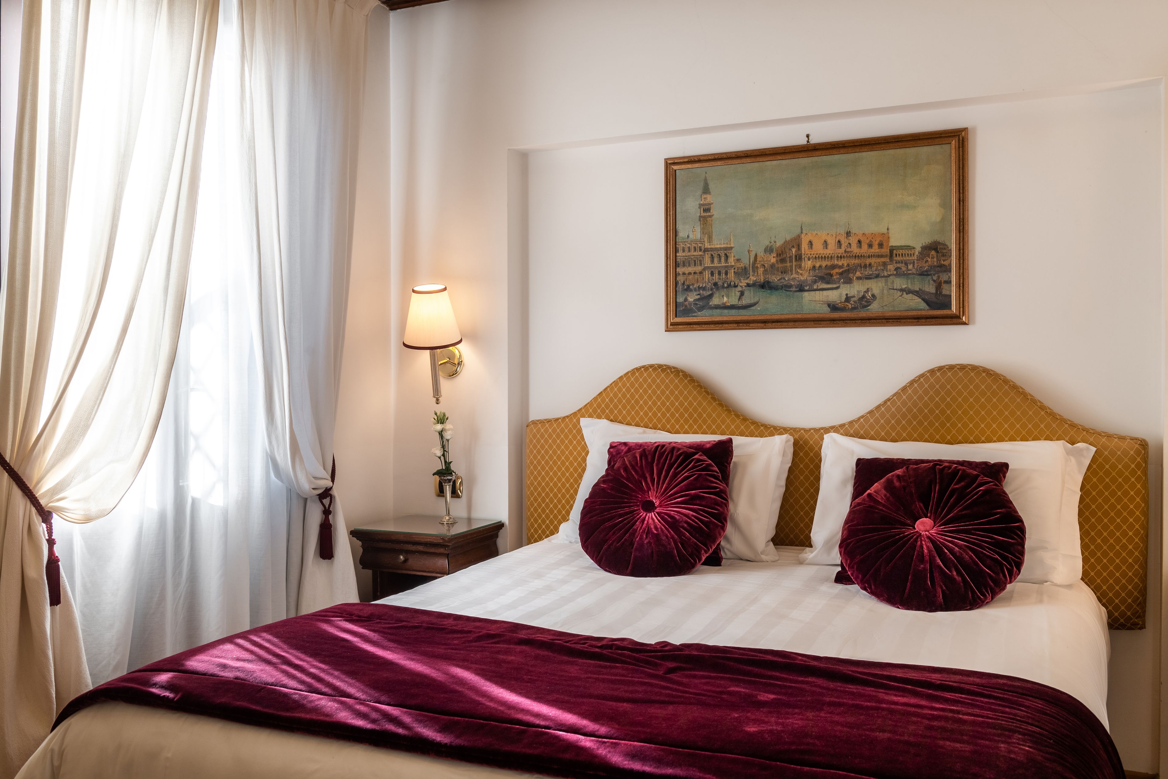 Ein ordentlich gemachtes Bett mit einer weißen und bordeauxroten Tagesdecke, zwei bordeauxroten Kissen und einem Kopfteil mit einem gemusterten Design. Ein Stadtbild hängt als Gemälde an der Wand über dem Bett.