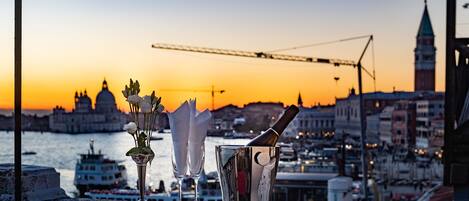 A balcony with a champagne bottle, glasses, and flowers, overlooking a cityscape at sunset.