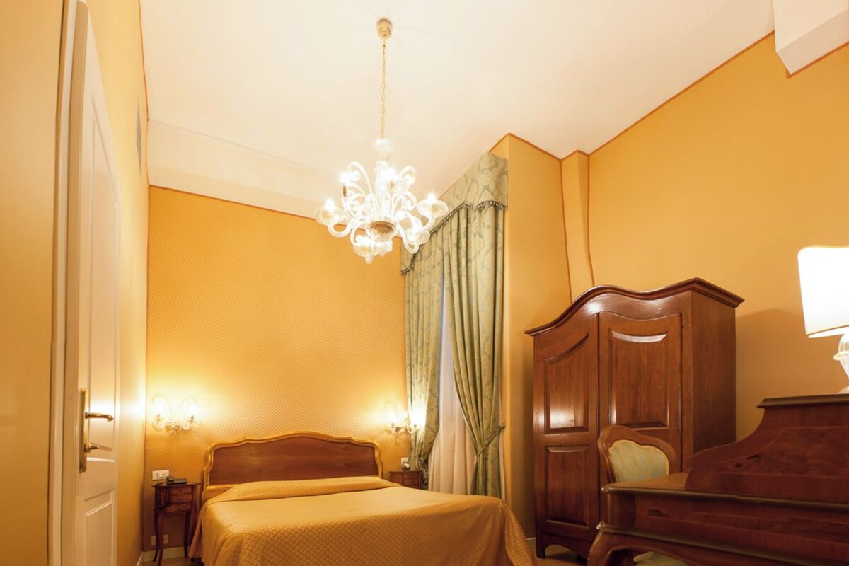 A hotel room with two beds, a wooden wardrobe, a desk with a lamp, and a chandelier.
