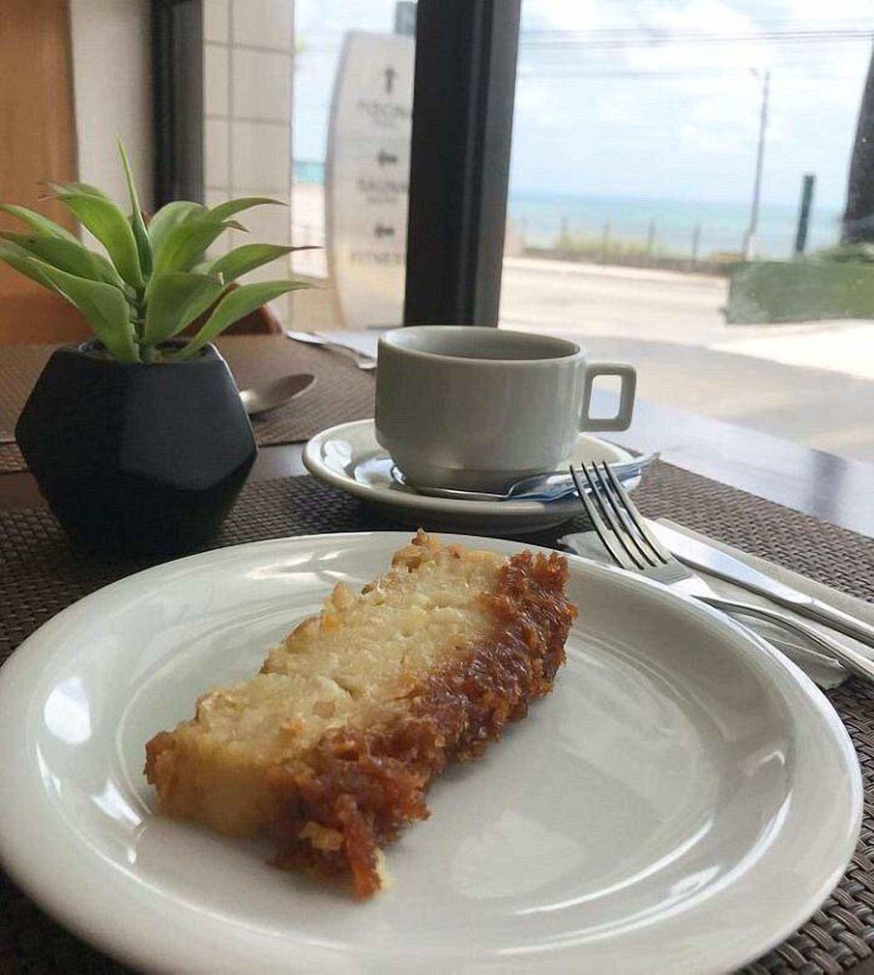 Un plato con un alimento, una taza, un tenedor y una planta en maceta sobre una mesa con vistas al mar.