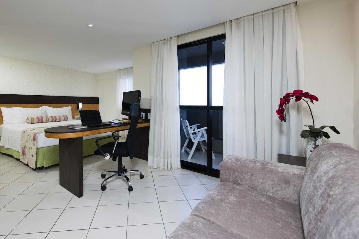 Habitación de hotel con cama, escritorio con computadora portátil, silla, sofá y ventana con cortinas.