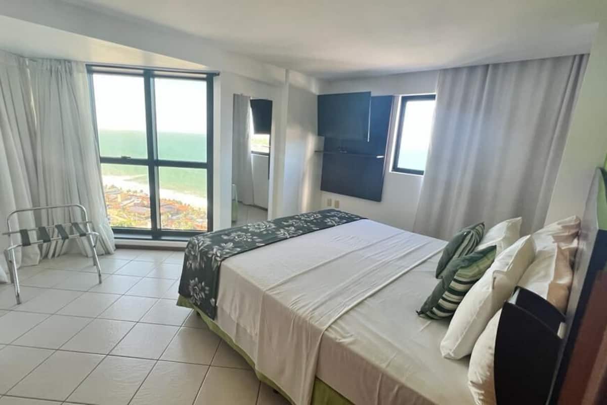 Habitación de hotel con cama, televisión, ventana con cortinas y vista al mar.