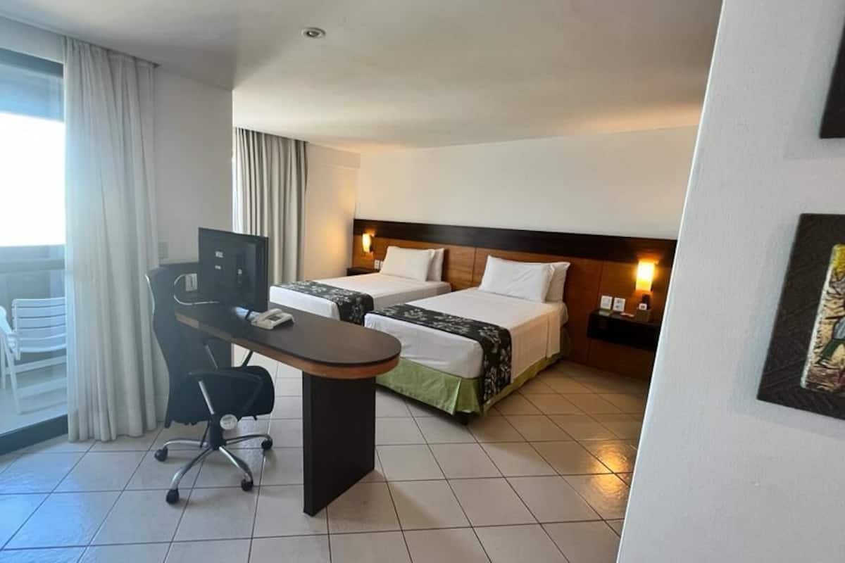 Una habitación de hotel con escritorio, dos camas, una silla y un ventanal grande.