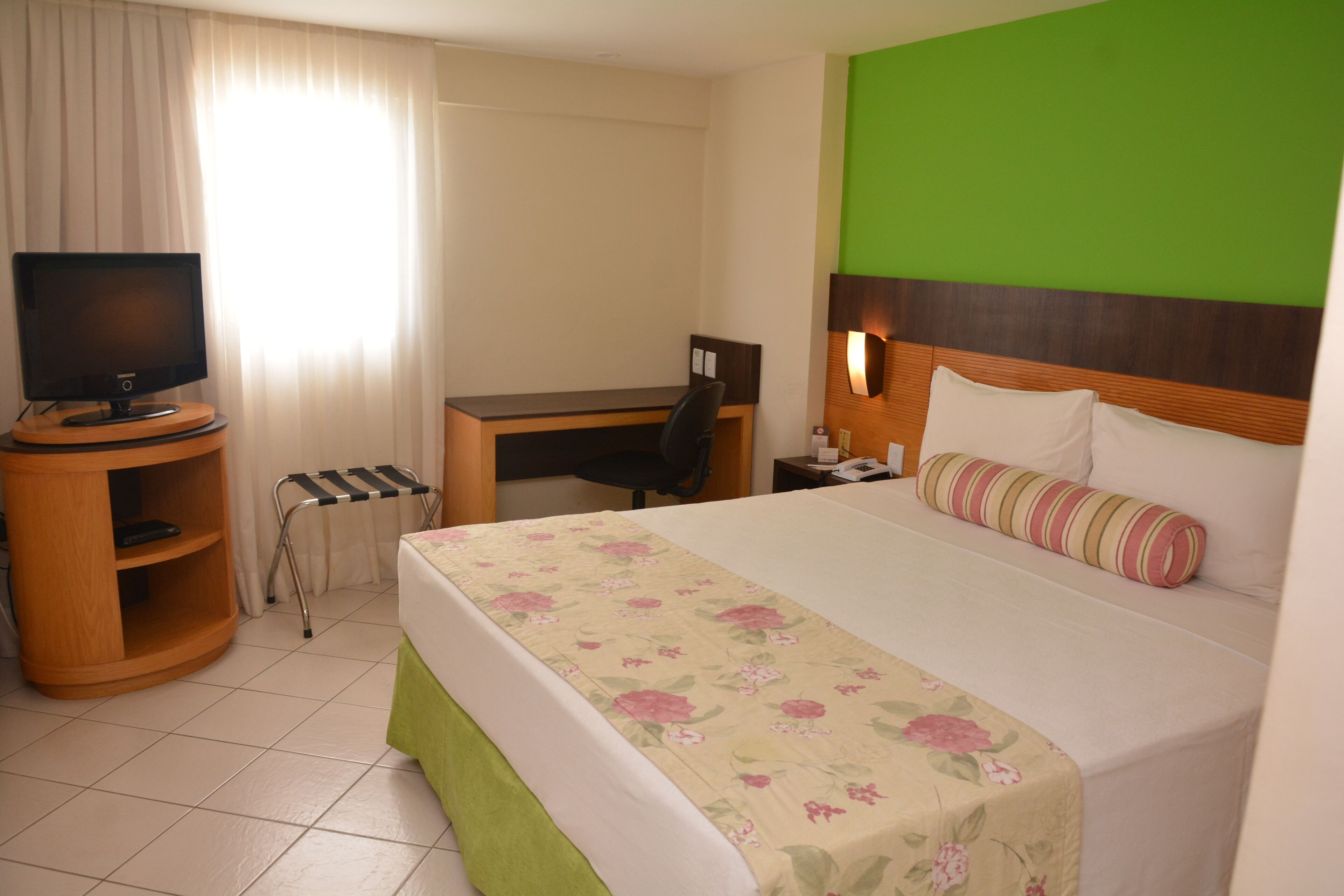 A hotel room with a bed, a desk, a chair, a television, and a small shelf.