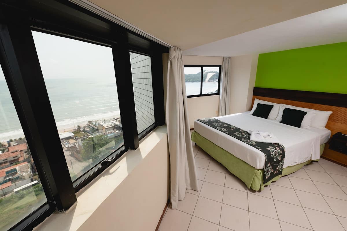 Habitación de hotel con un ventanal que ofrece vistas al mar, una cama bien hecha y una pared de acento verde.