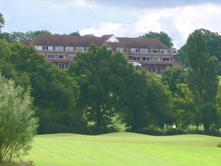 A large, multi-story building with a symmetrical roofline, surrounded by lush greenery and a well-maintained lawn.