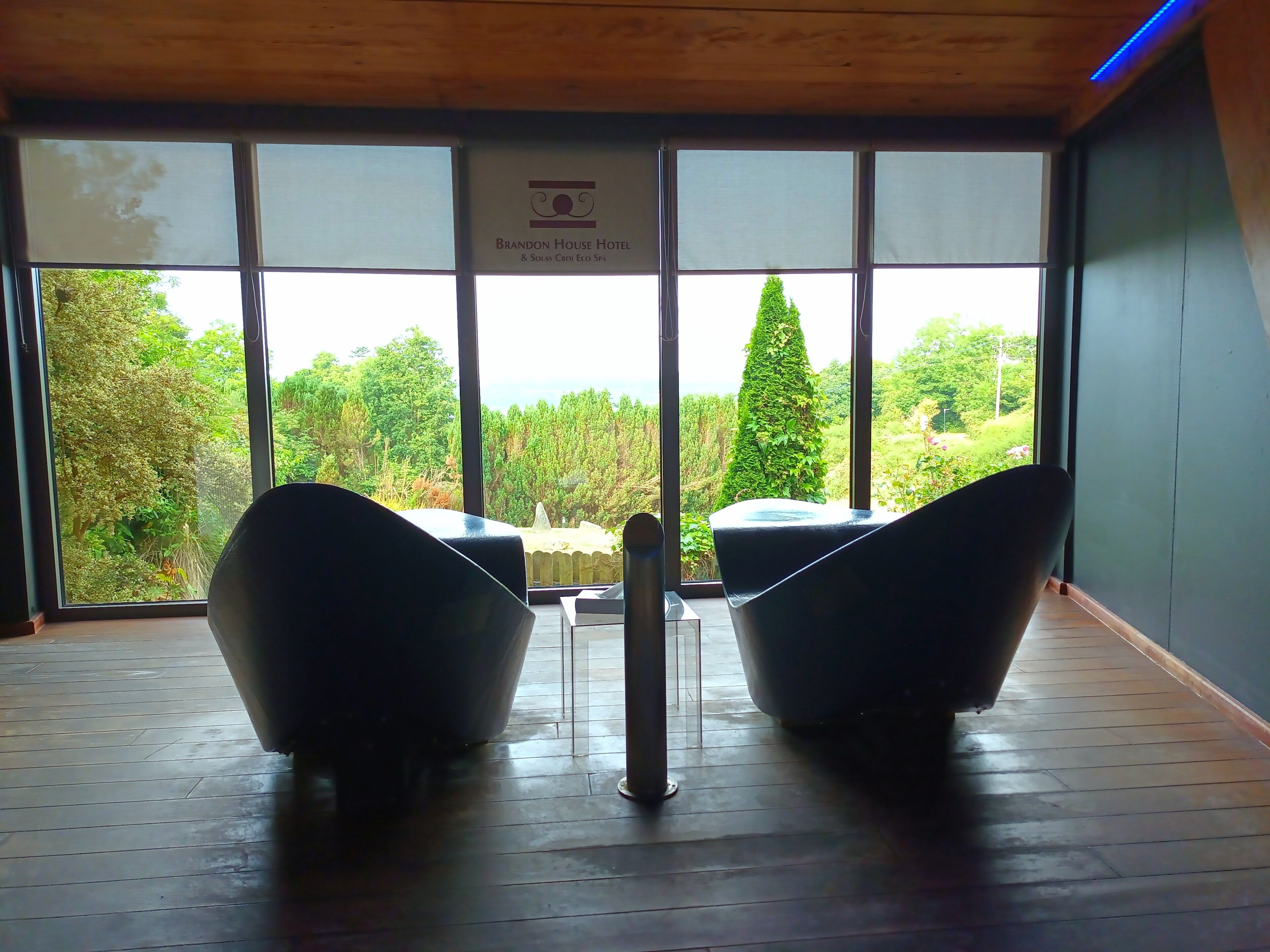 A room with a large window, two black chairs, a small table, and a sign reading "Brandon House Hotel & Solas Croi Eco Spa".