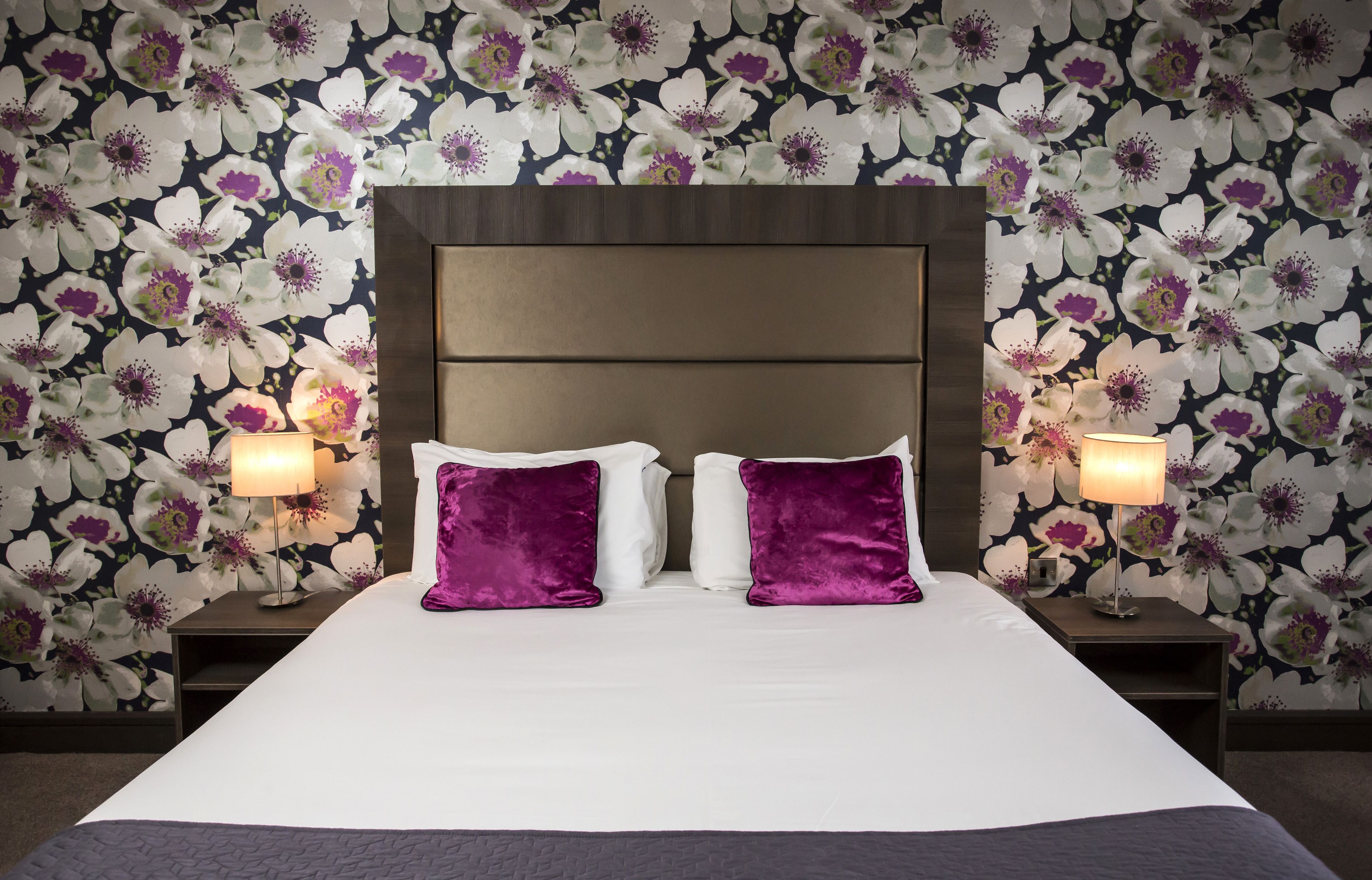 Une chambre d’hôtel avec un grand lit, deux oreillers violets, une tête de lit en bois et un papier peint à motifs floraux.