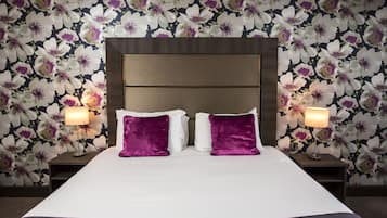 Habitación de hotel con una cama grande, dos almohadas moradas, cabecera de madera y papel tapiz con motivos florales.