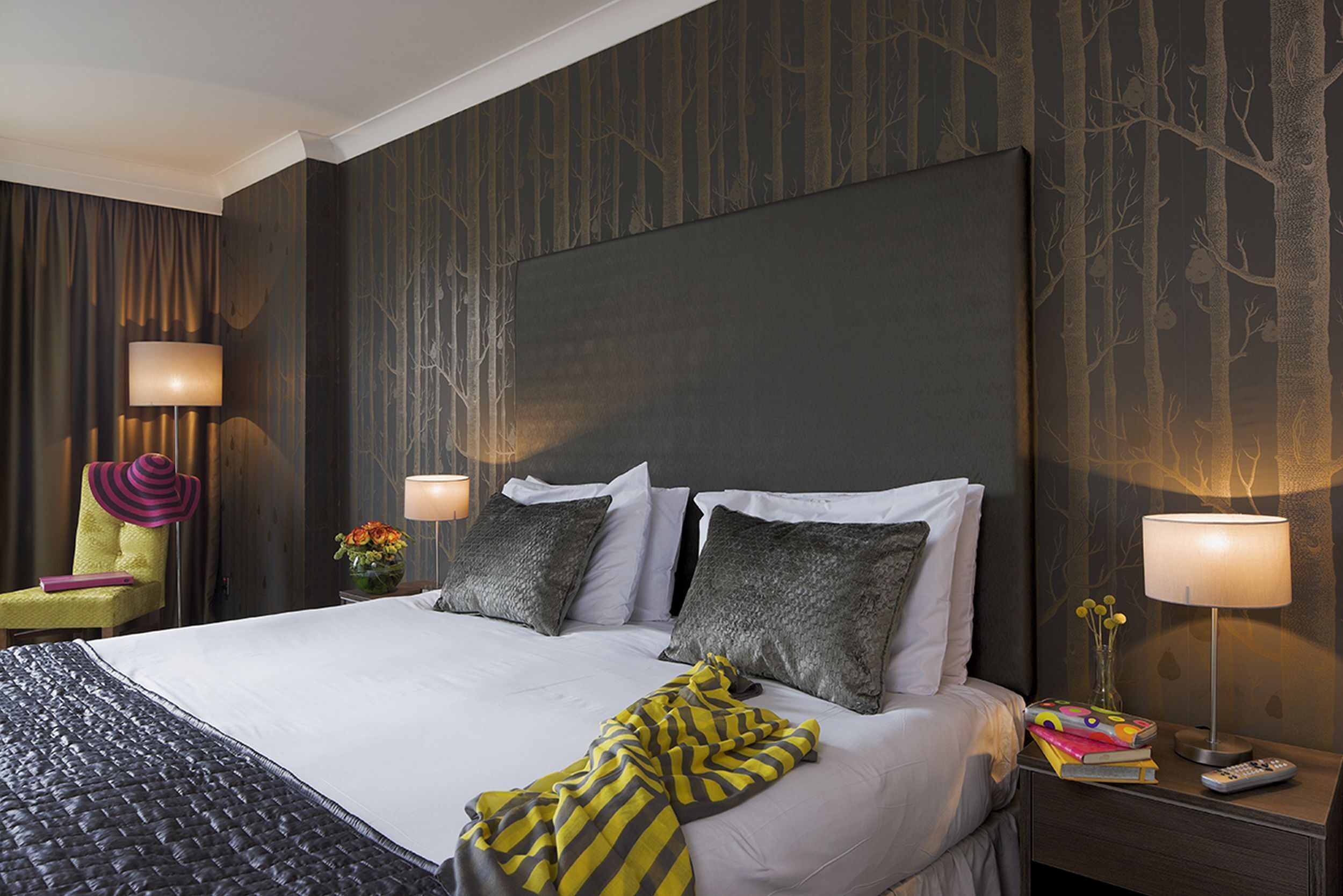 A hotel room with a large bed, two bedside tables, a chair, and a wall with a tree pattern.