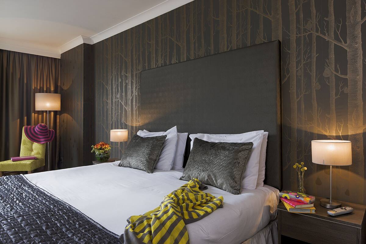 Habitación de hotel con una cama grande, dos mesitas de noche, una silla y una pared con diseño de árbol.