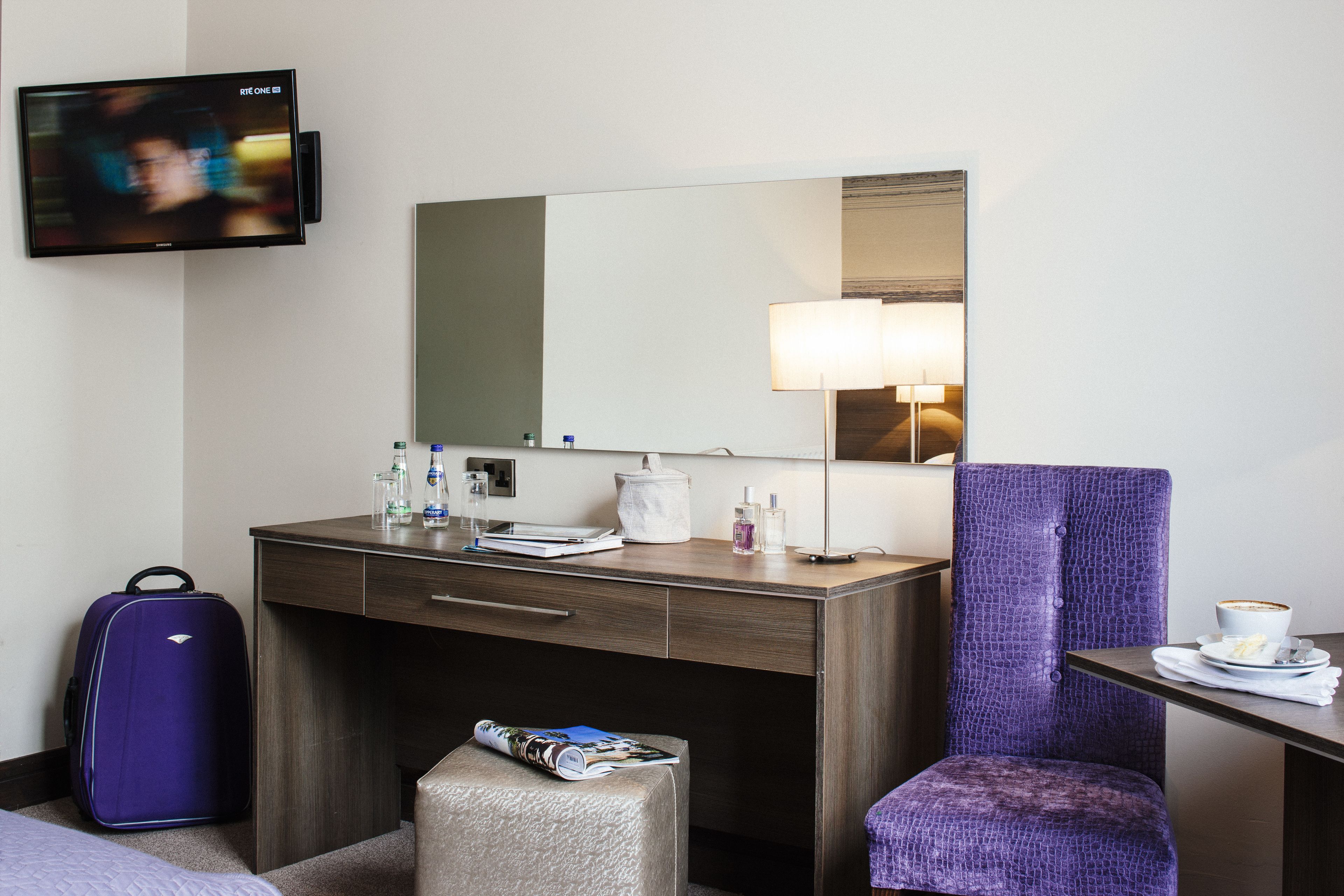 Habitació d'hotel amb escriptori de fusta, una cadira morada, una taula petita amb una tassa, una maleta, una revista i un televisor fixat a la paret.