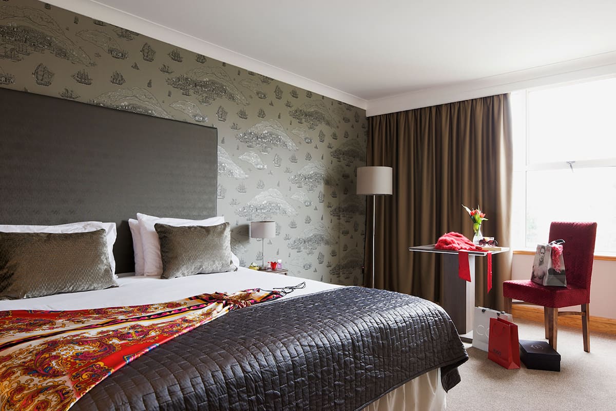 A hotel room with a large bed, a desk with a lamp, a chair, and a window with curtains.