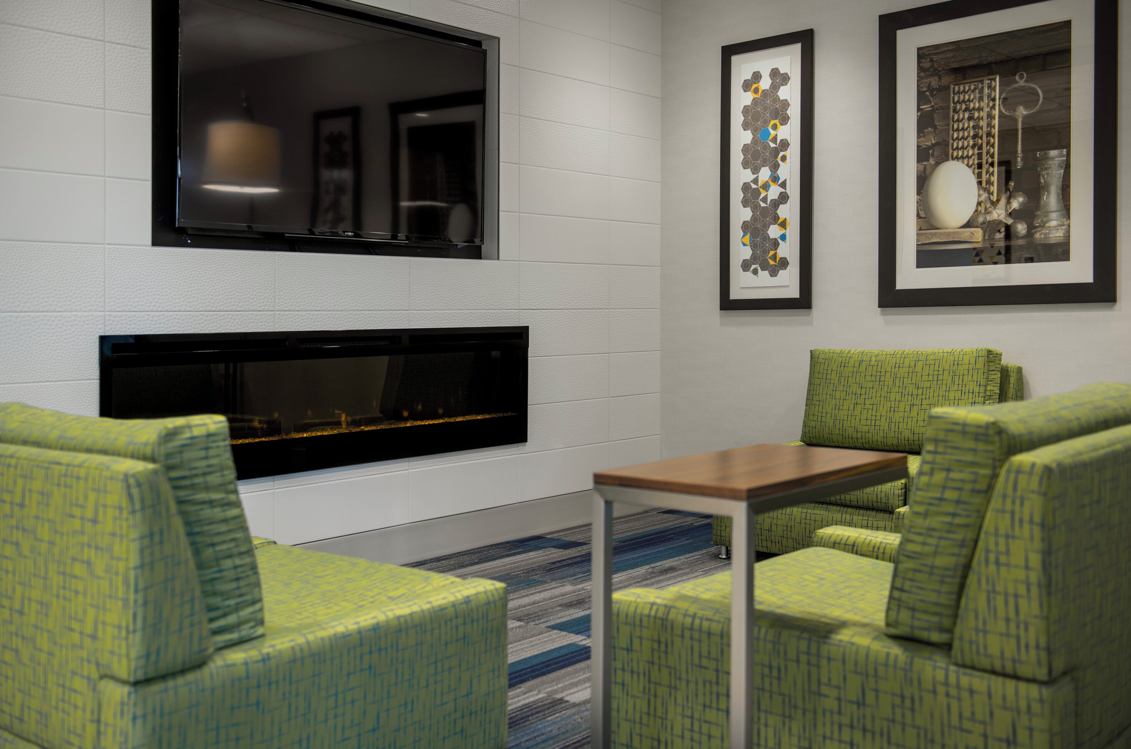 A modern lounge area with a fireplace, green patterned chairs, a small table, and framed artwork on the wall.
