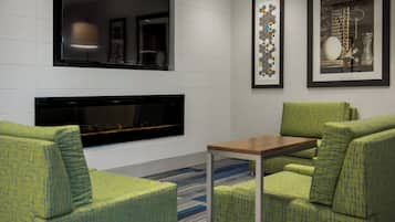 A modern lounge area with a fireplace, green patterned chairs, a small table, and framed artwork on the wall.