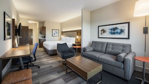 A hotel room with a bed, a gray sofa, a wooden coffee table, a desk with a chair, and a small table with a lamp.