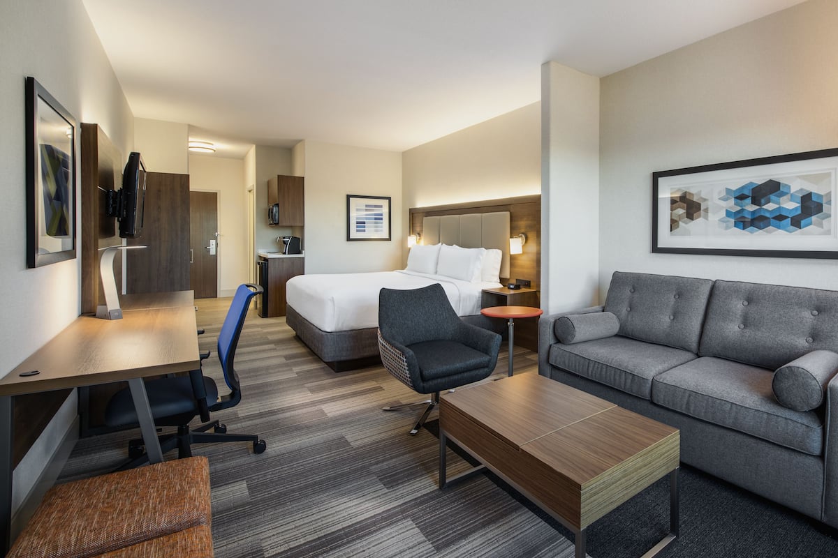 A hotel room with a bed, a gray sofa, a wooden coffee table, a desk with a chair, and a small table with a lamp.