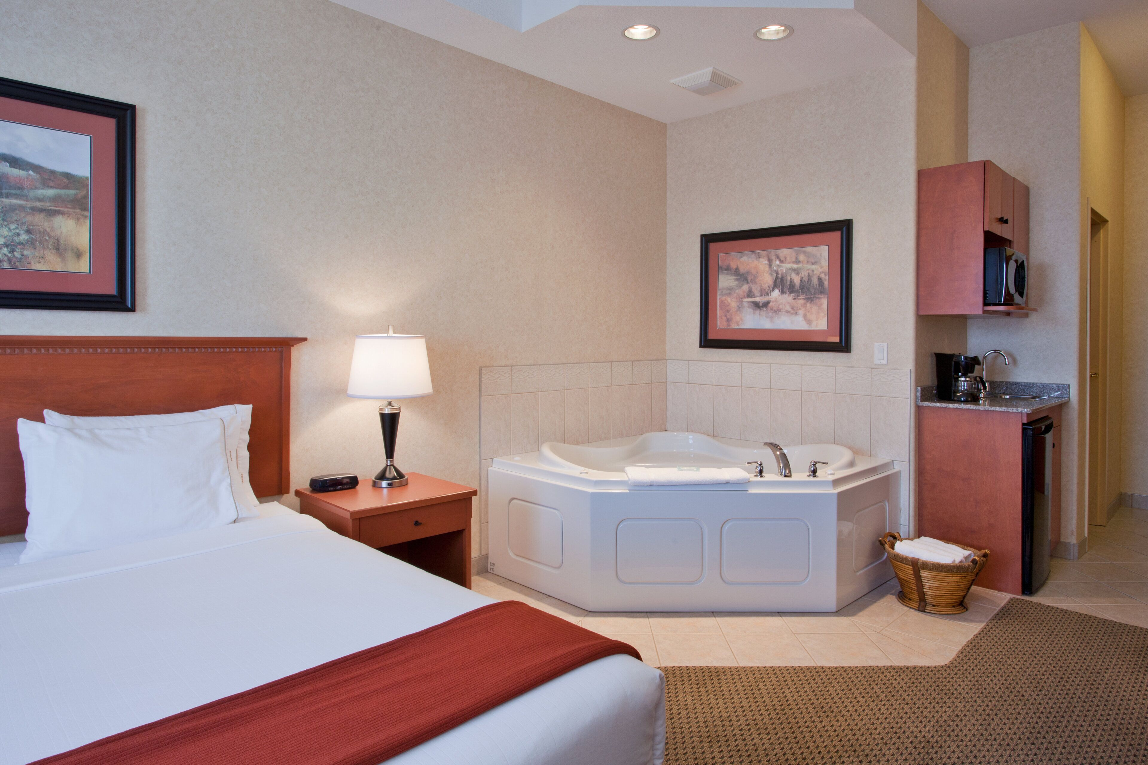 A hotel room with a large bed, a sitting area with a lamp, a bathtub, and a small kitchenette.
