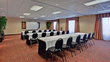 Un salón de conferencias con varias mesas y sillas, una pantalla para proyector y una planta grande.