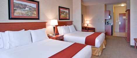 A hotel room with two beds, a desk, a chair, a lamp, a small table, and a door leading to a kitchenette.
