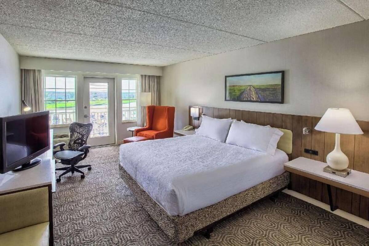 A hotel room with a bed, a desk with a TV, a chair, a nightstand with a lamp, and a painting on the wall.