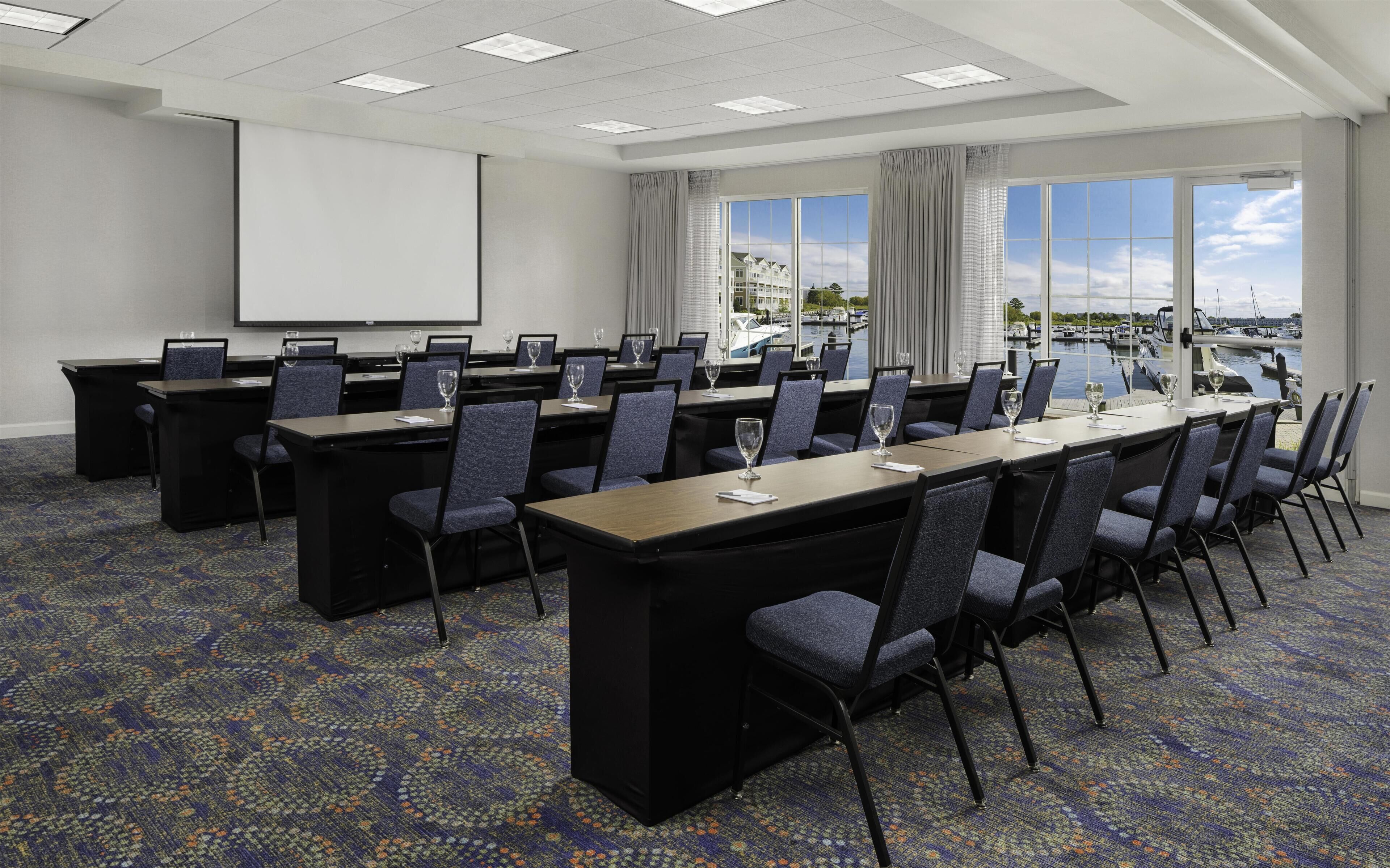 A conference room with a large screen, multiple tables, and chairs, overlooking a marina with boats.