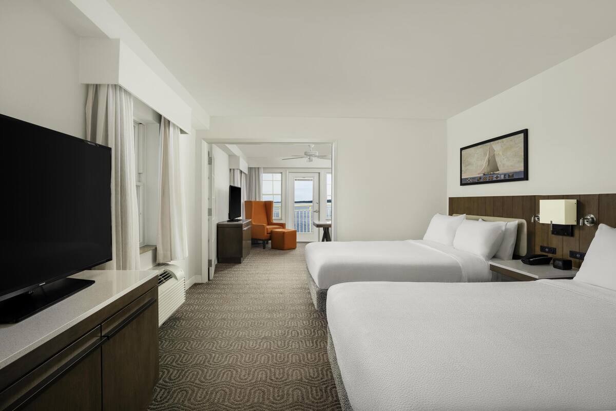 A hotel room with two beds, a TV, a desk, and a view of the sea.