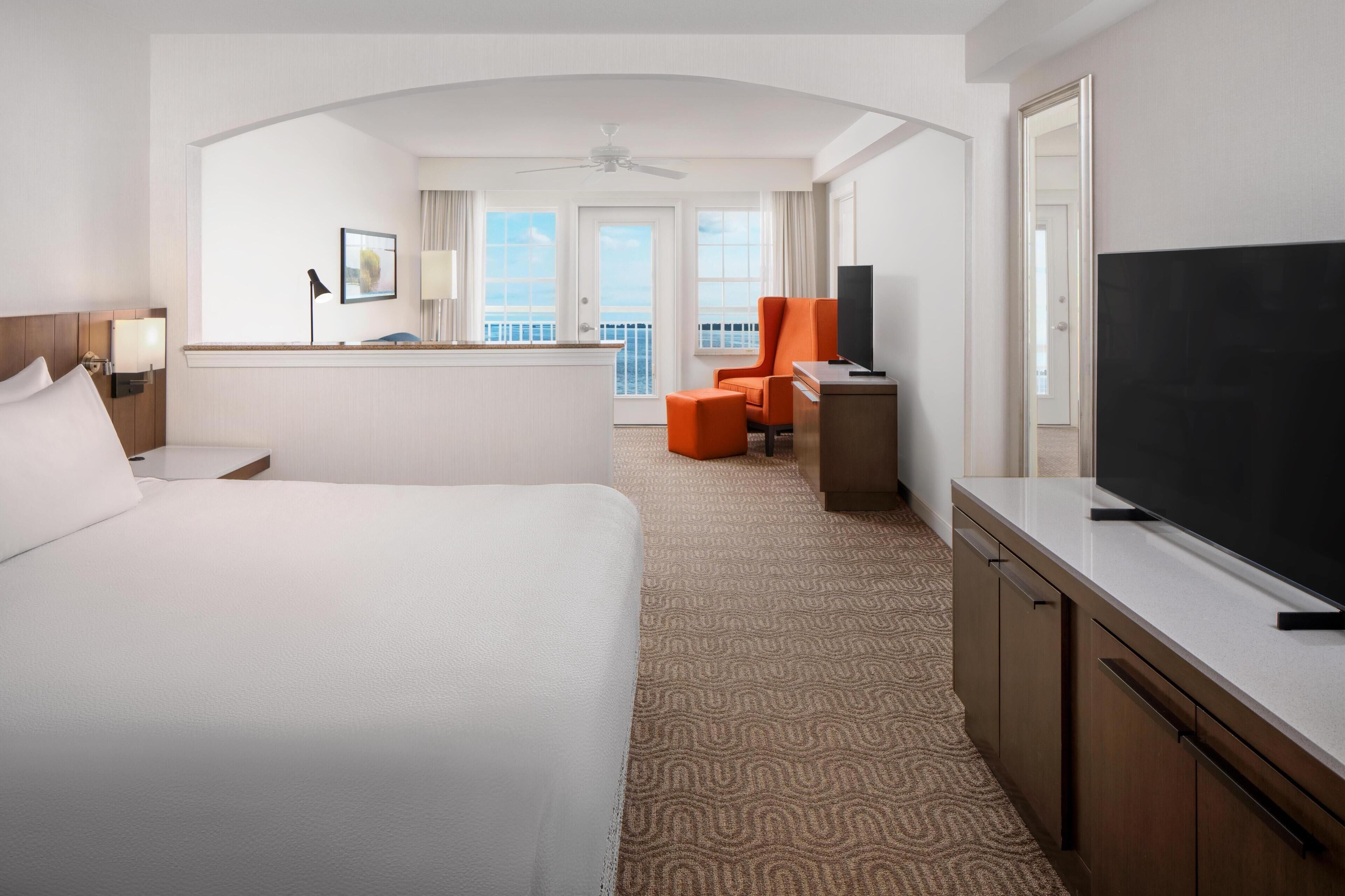 A hotel room with a large bed, a desk with a flat-screen TV, an orange chair, and a balcony with a view.