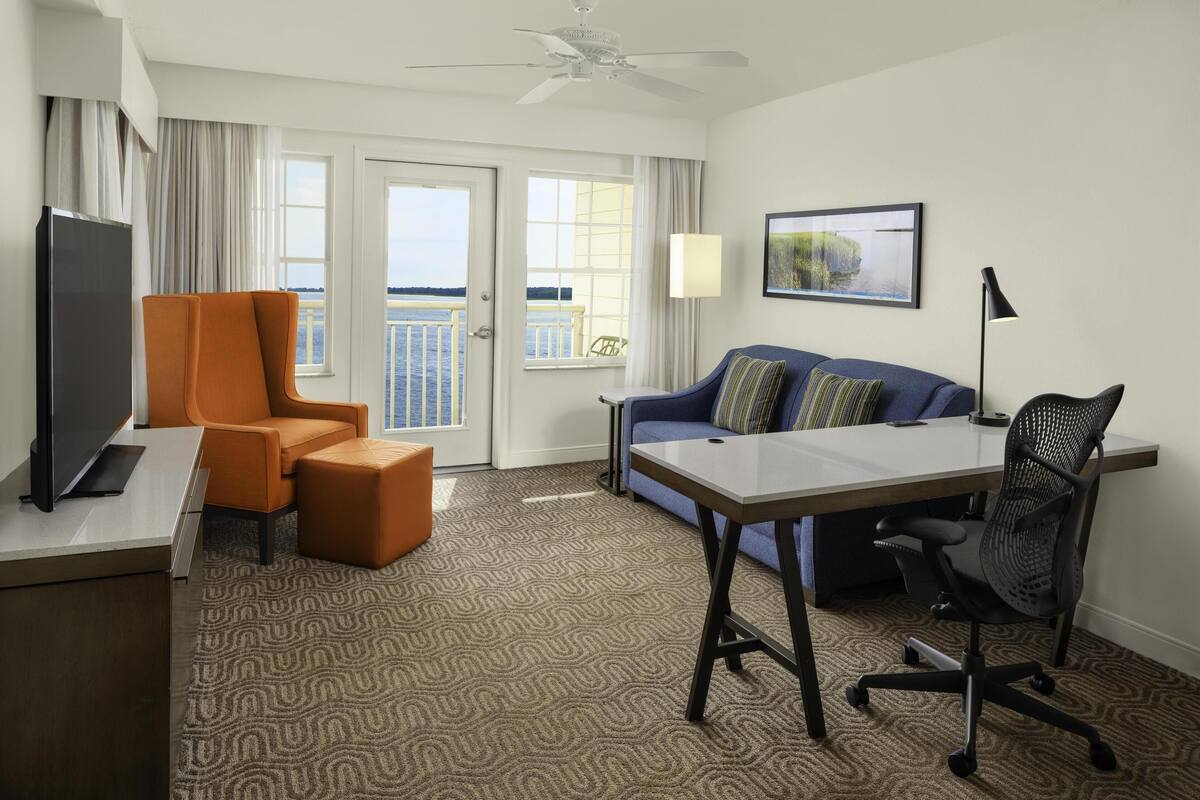 A hotel room with a desk, a television, an orange chair, a blue sofa, a black office chair, and a ceiling fan.