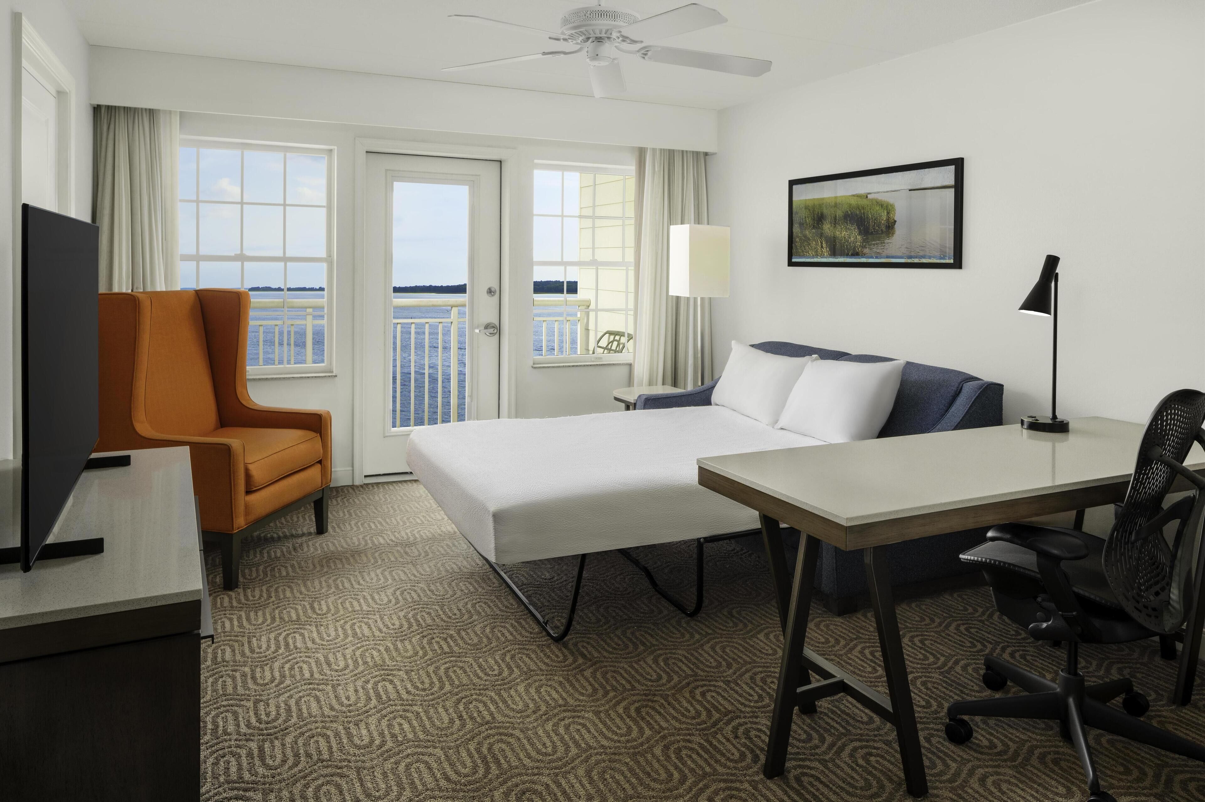 Habitación de hotel con cama, escritorio, silla, televisión, ventilador de techo y vista al agua.
