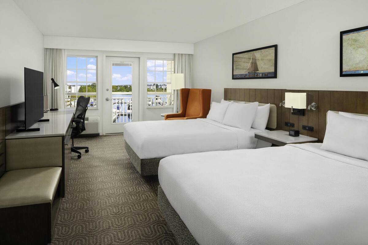 A hotel room with two beds, a desk, a chair, and a TV. There is a balcony with a view of the water and buildings.