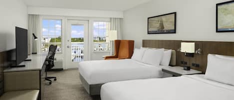 Habitación de hotel con dos camas, un escritorio, una silla y un televisor. Tiene un balcón con vistas al agua y edificios.