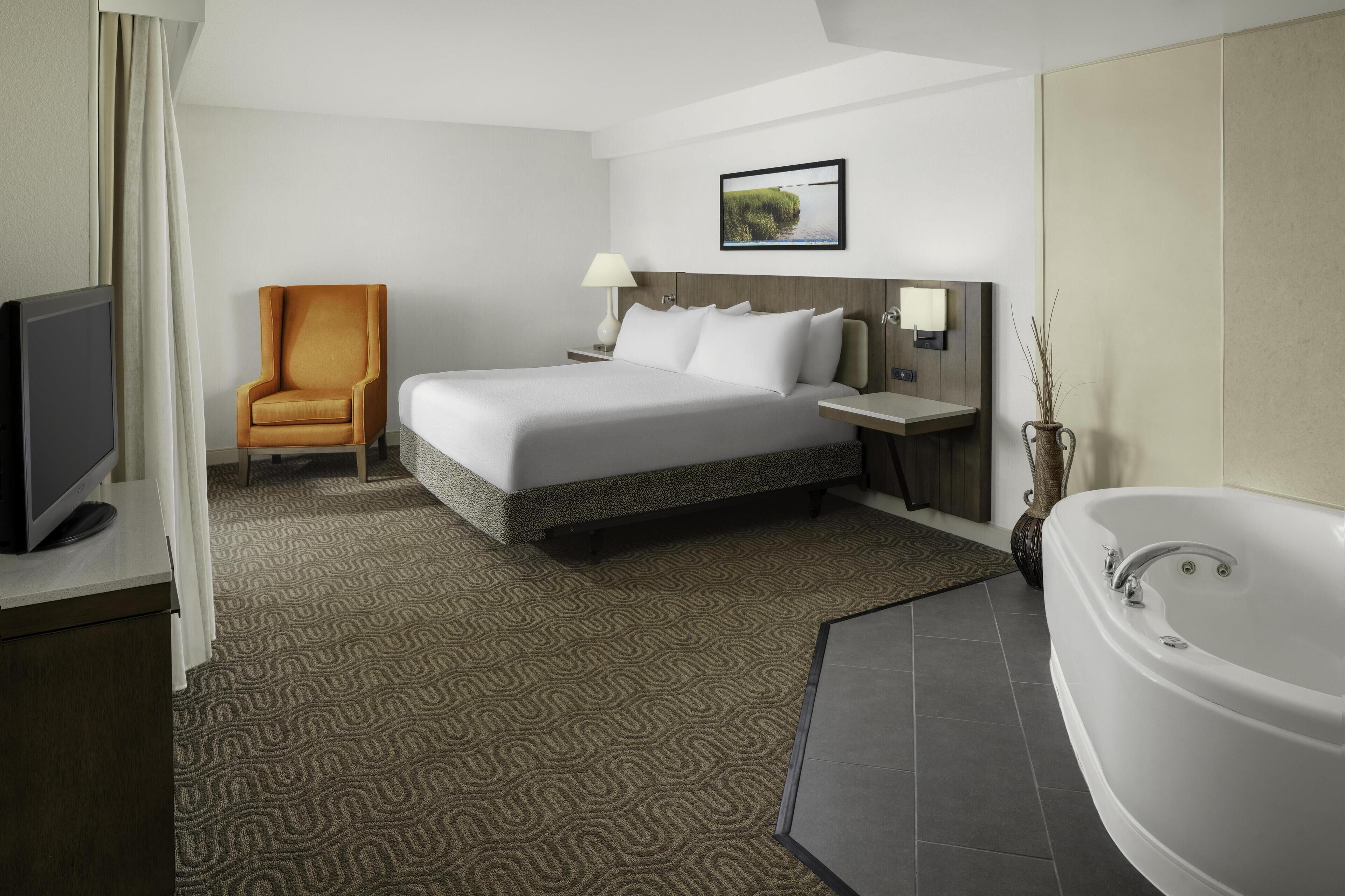 A hotel room with a large bed, a chair, a TV, a desk, a lamp, a picture on the wall, and a bathtub.
