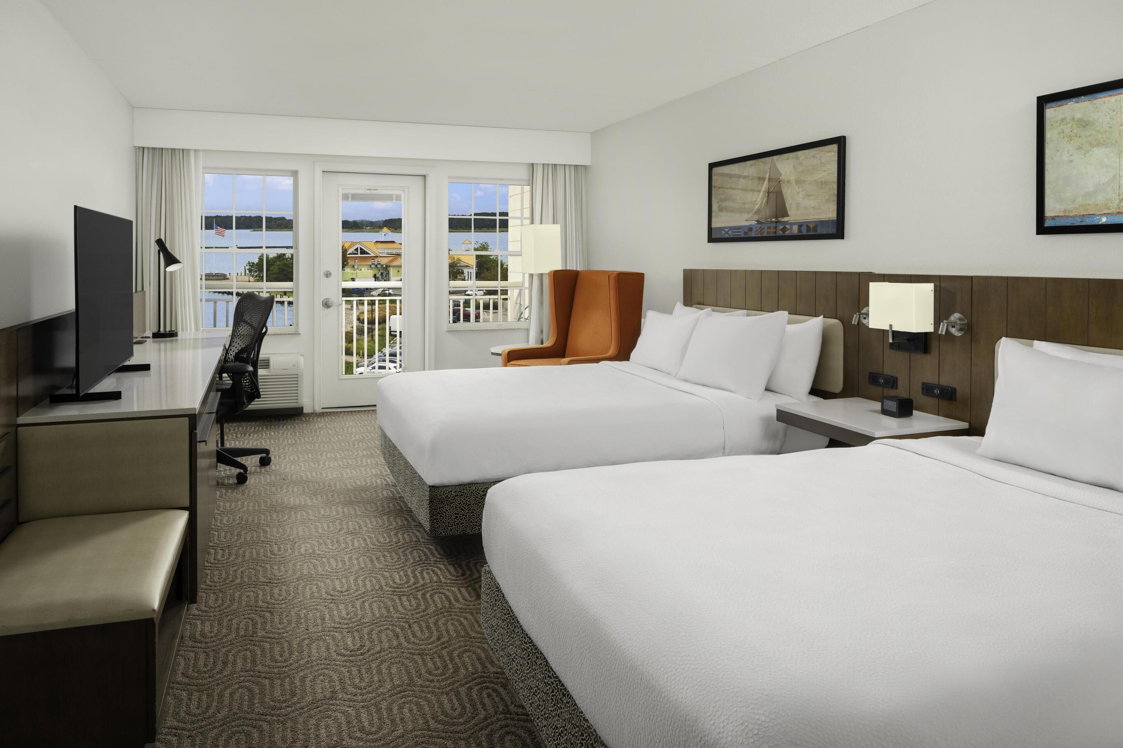 A hotel room with two beds, a desk, a TV, and a view of the outdoors.
