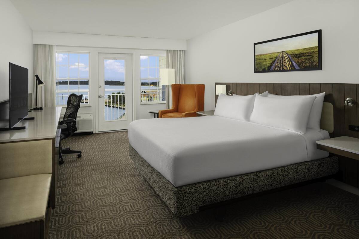 A hotel room with a large bed, a desk with a chair, a TV, and a view of the outdoors through a sliding door.