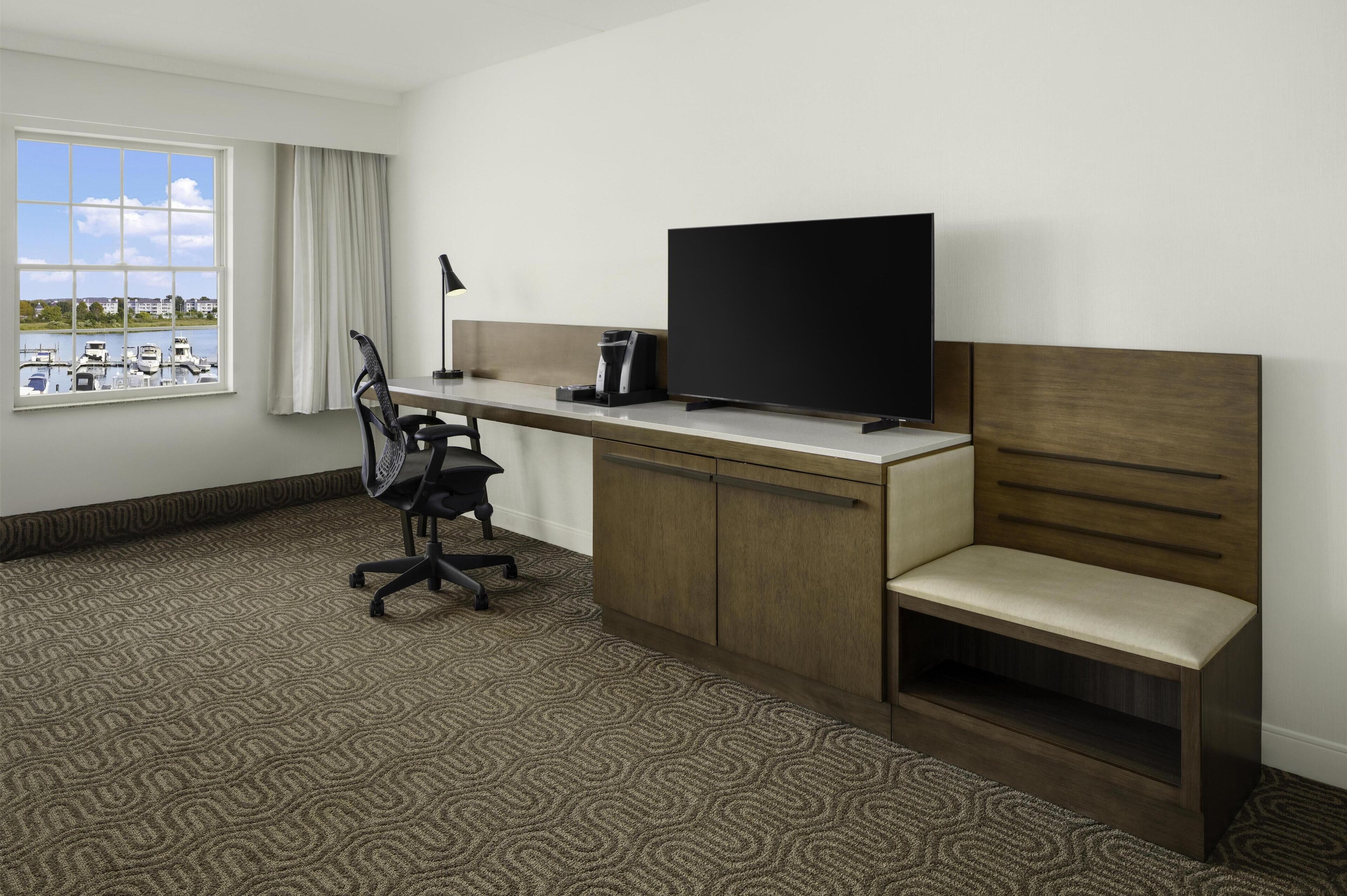 A hotel room with a large window, a desk with a chair, a TV, and a bench.
