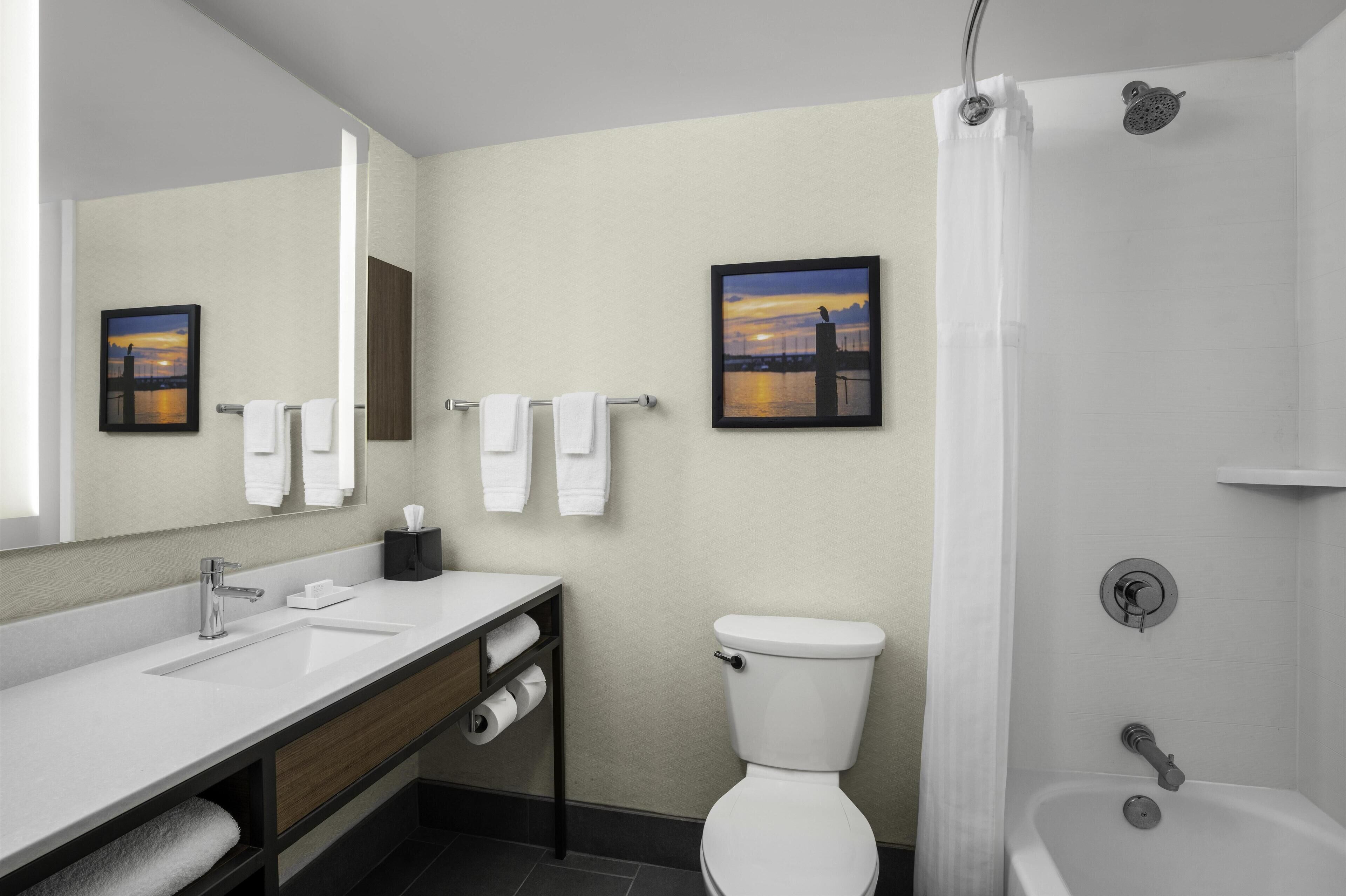 A modern bathroom with a white sink, vanity, and toilet. There are two framed pictures on the wall, a shower curtain, and a bathtub.