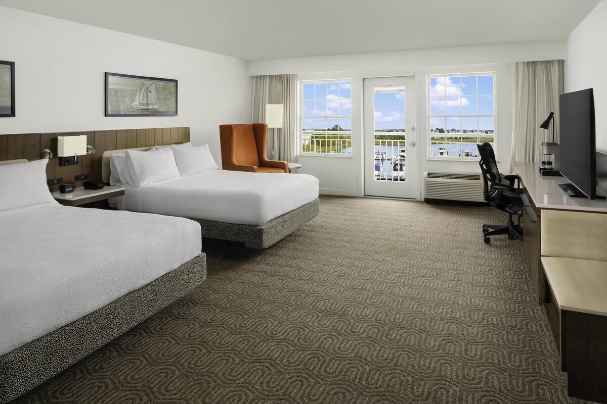 A hotel room with two beds, a desk, a TV, and a balcony with a view of the water.