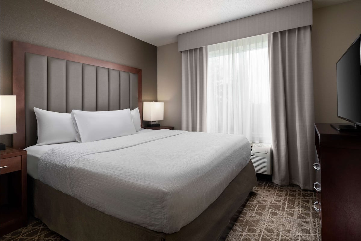 A hotel room with a large bed, a headboard, two bedside tables with lamps, a television, and a window with curtains.