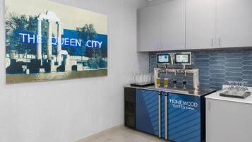 A hotel bar area with a large 'THE QUEEN CITY' sign, a minibar, and a digital screen displaying beverage options.