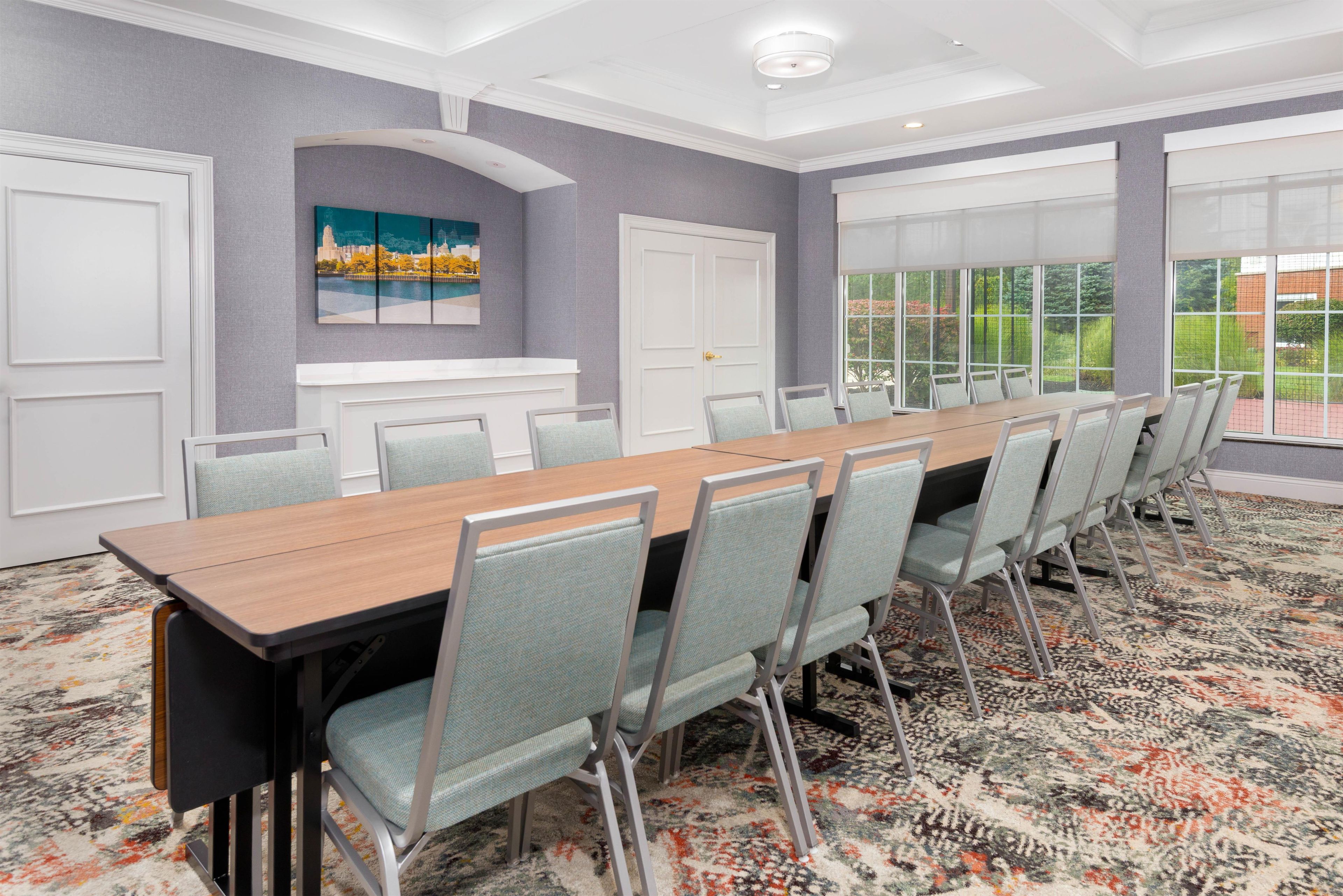 A conference room with a long table, chairs, and a view of greenery through large windows.