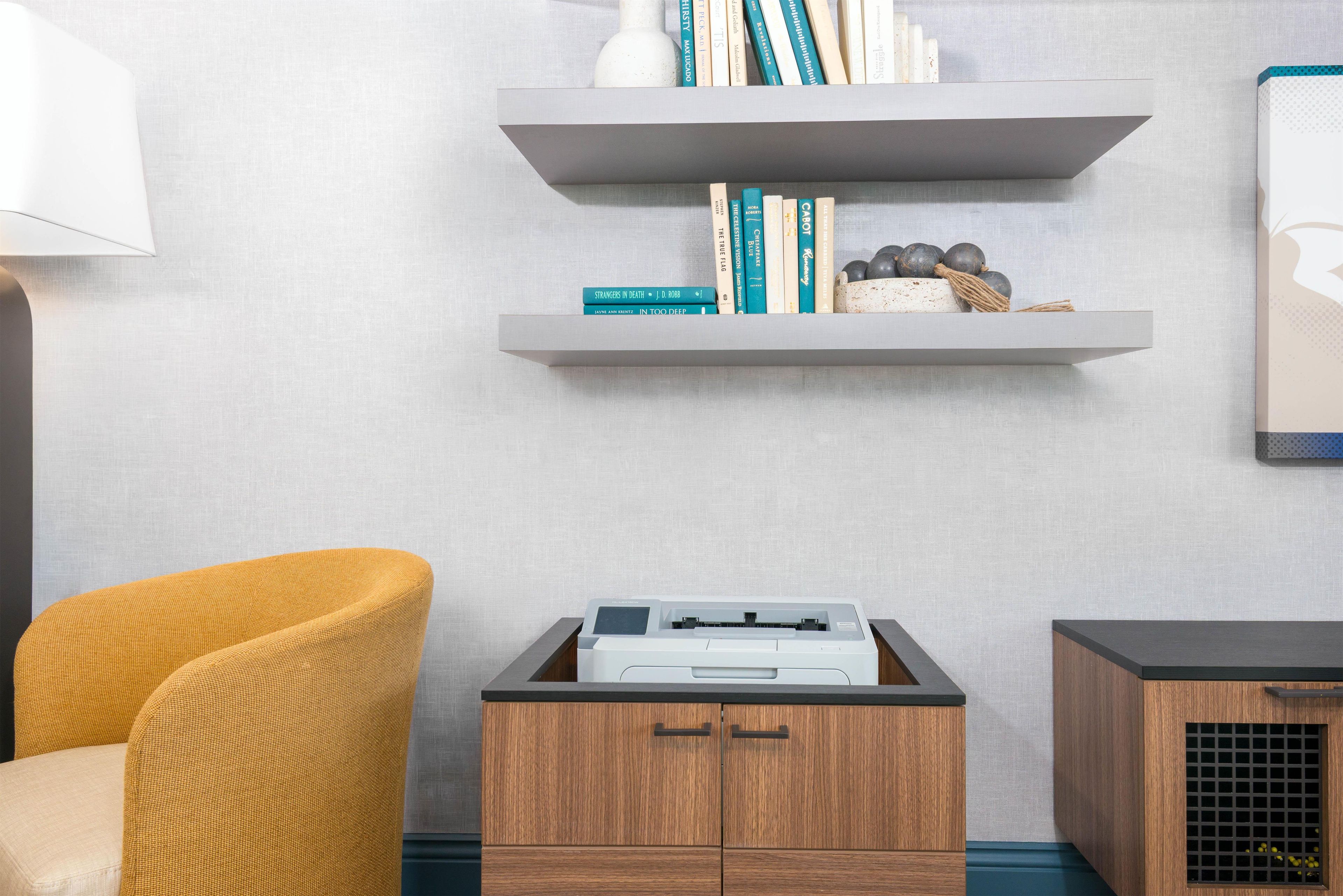 A room with a printer on a wooden cabinet, a yellow chair, and two wall-mounted shelves with books and decorative items.