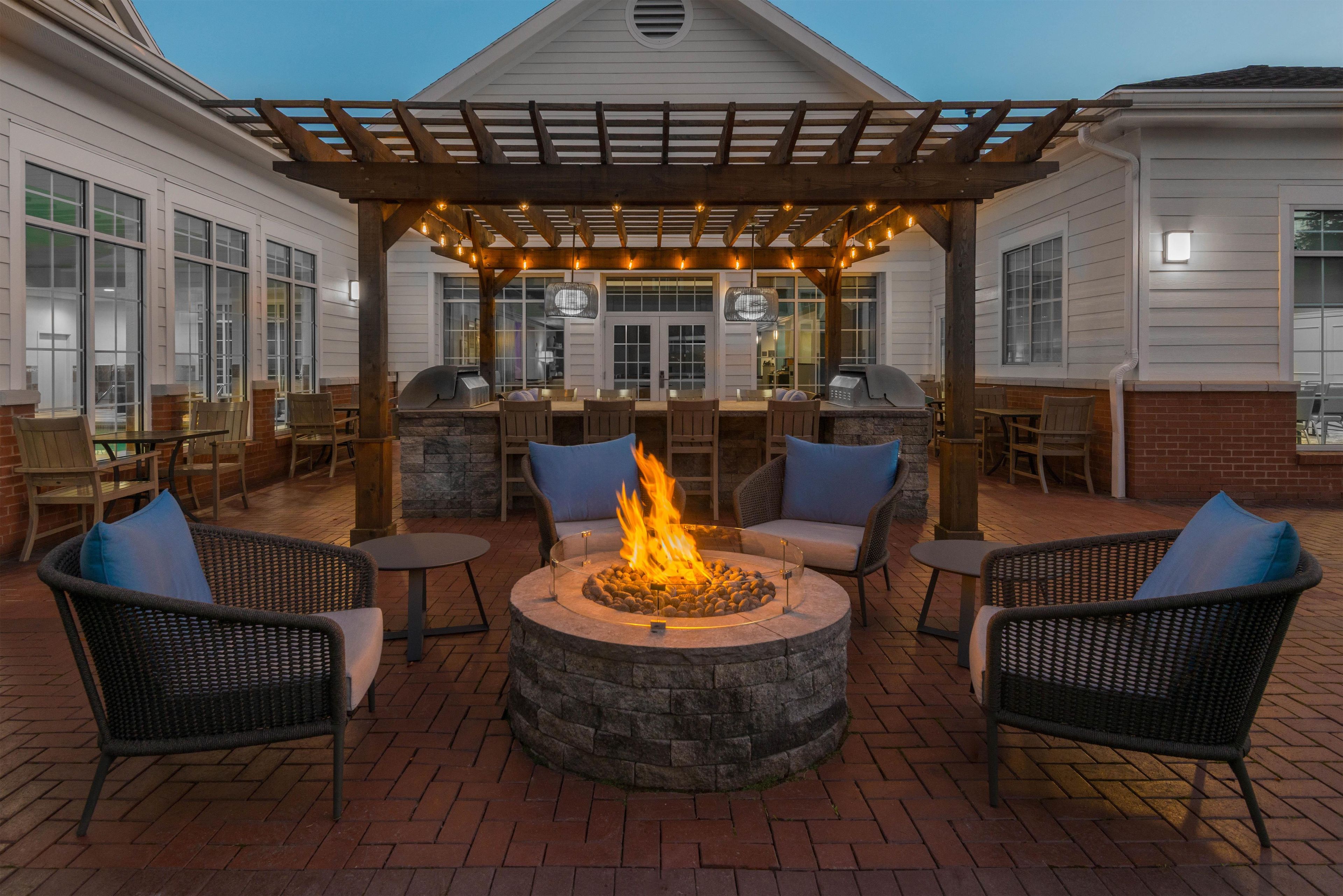 An outdoor patio area with a stone fire pit, wicker chairs, and a pergola with string lights.