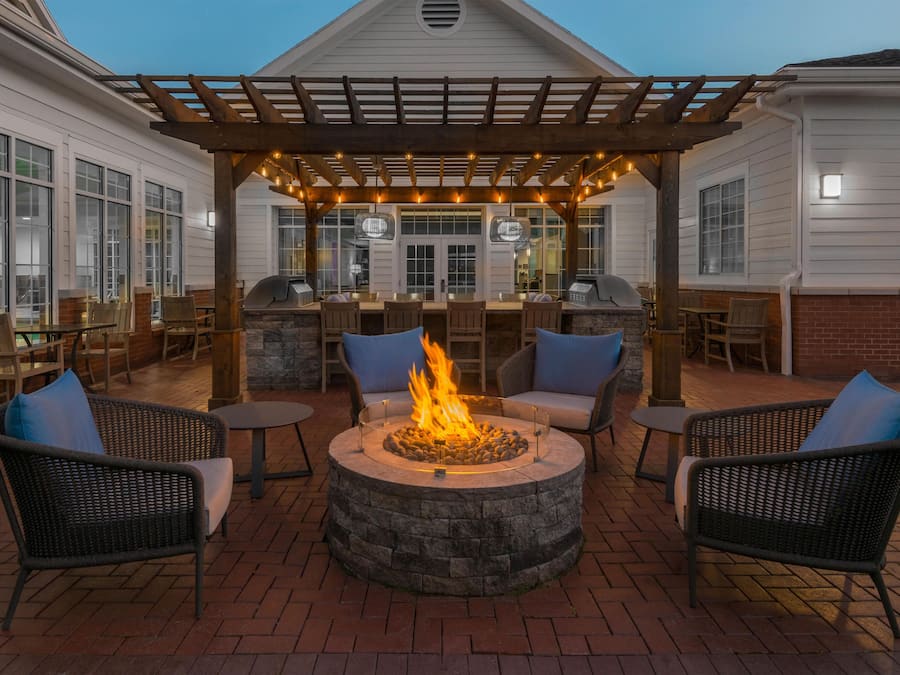 An outdoor patio area with a stone fire pit, wicker chairs, and a pergola with string lights.