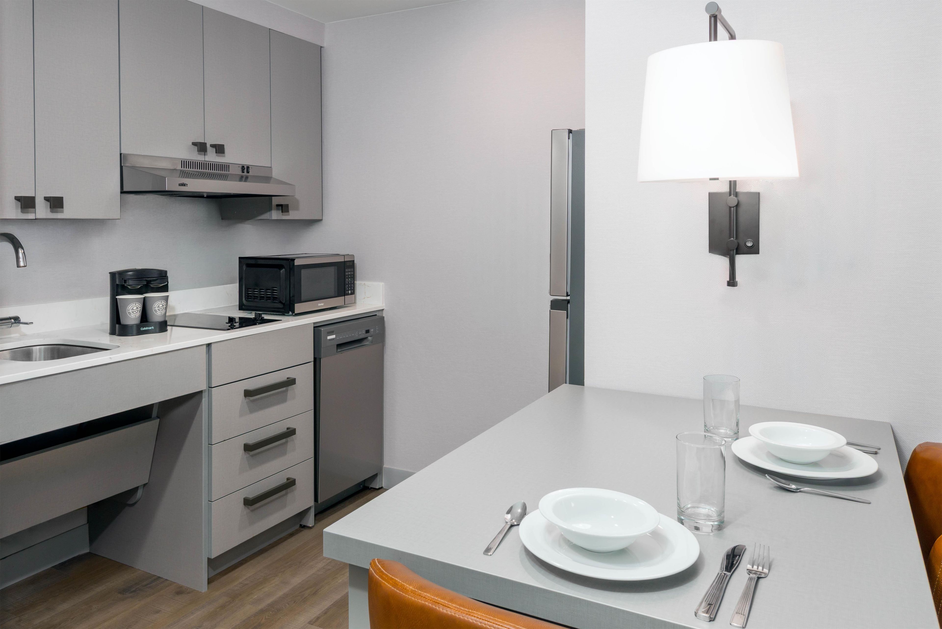 A modern kitchen with a built-in dishwasher, a microwave, and a coffee maker. There is a dining table with plates, glasses, and cutlery.