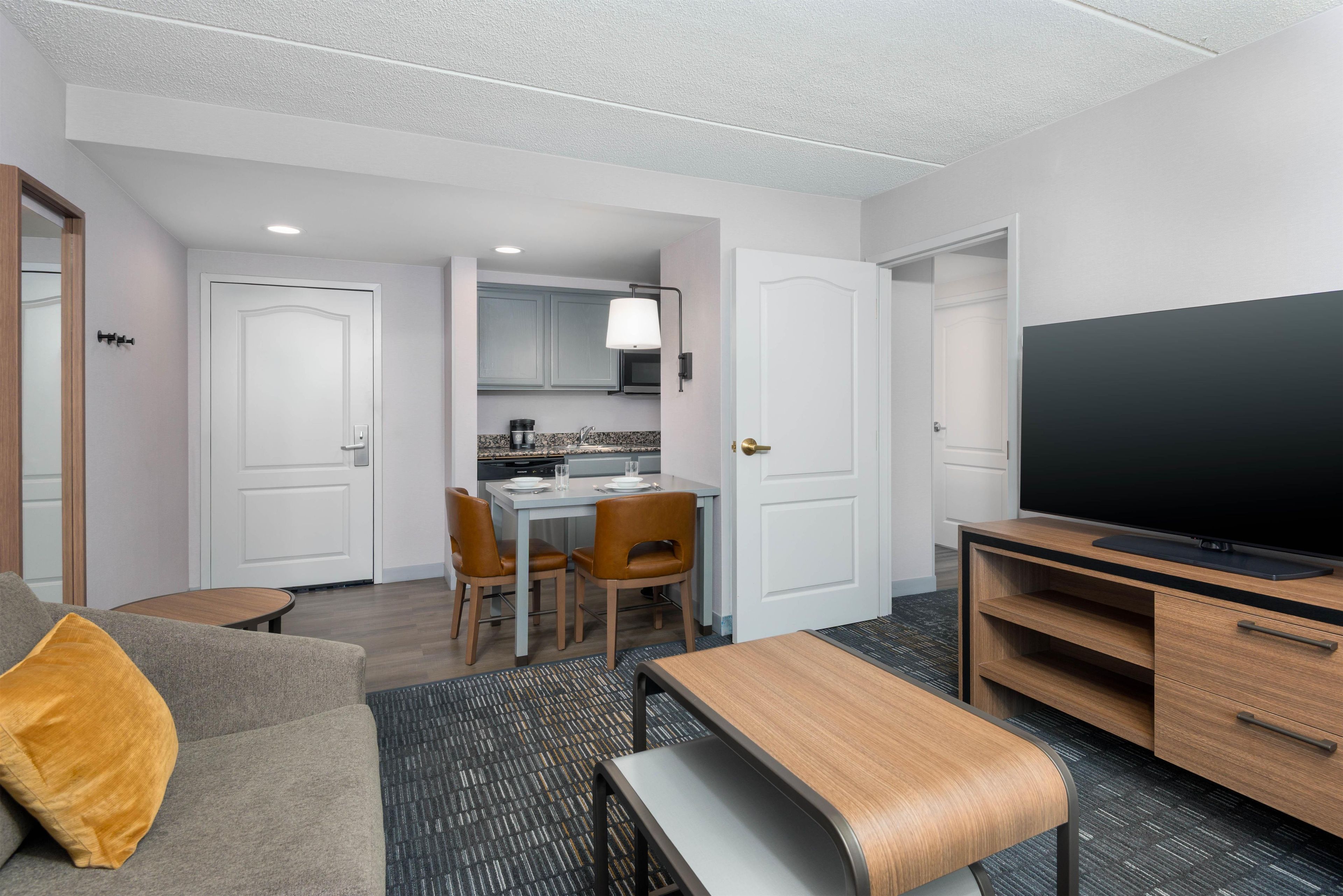 A modern hotel room with a sofa, a TV, a dining area with chairs and a table, and a kitchen area.