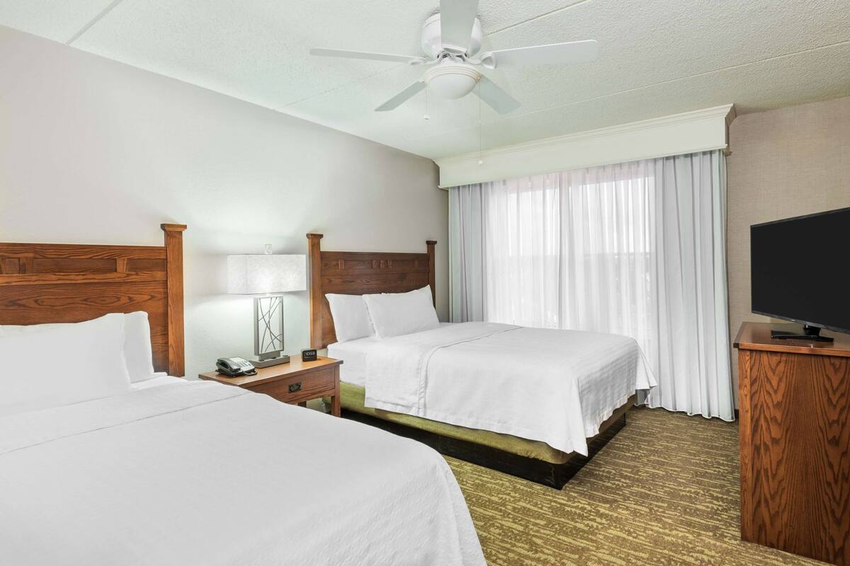 A hotel room with two beds, a ceiling fan, a TV, and a window with curtains.