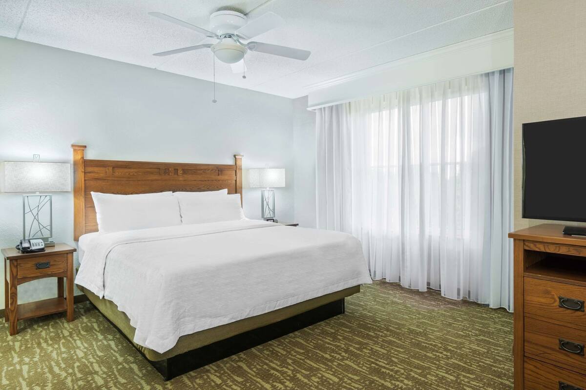 A hotel room with a bed, a ceiling fan, a TV, a nightstand with a lamp, and a window with curtains.
