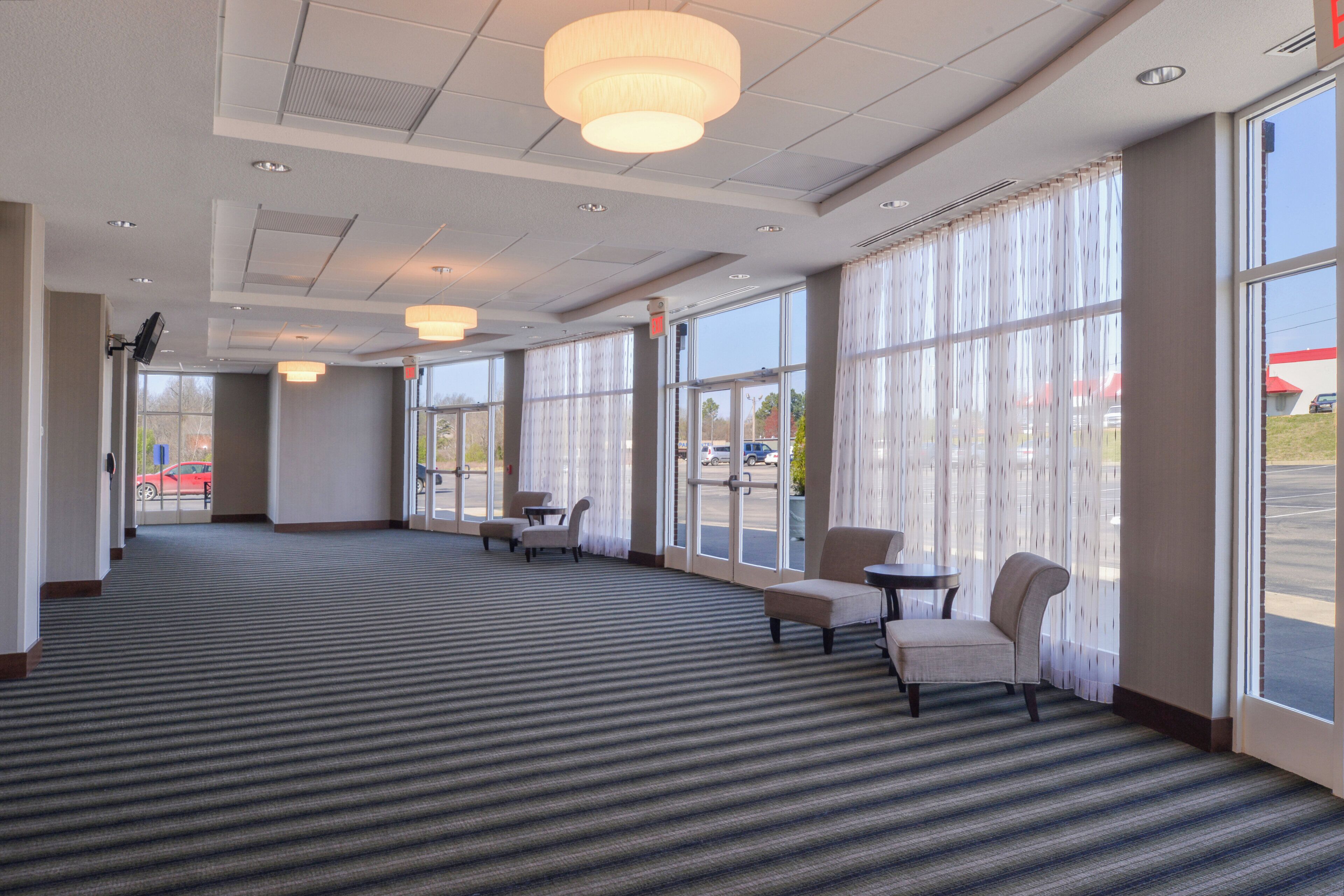 A hotel lobby with a carpeted floor, seating area with chairs, large windows, and ceiling lights.