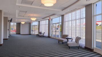 A hotel lobby with a carpeted floor, seating area with chairs, large windows, and ceiling lights.